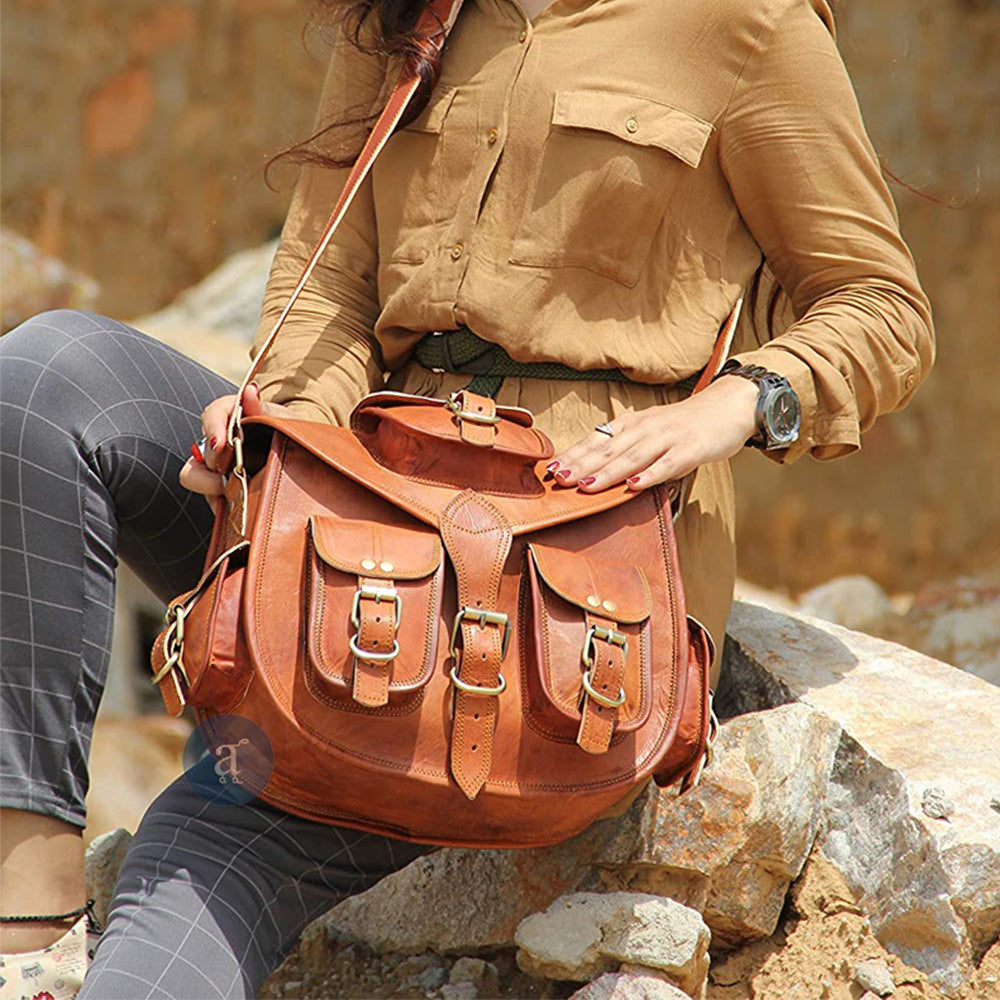 A Woman Sitting On Rocks, Holding A Large Tote With Multiple Buckles, Wearing A Brown Shirt And Plaid Pants